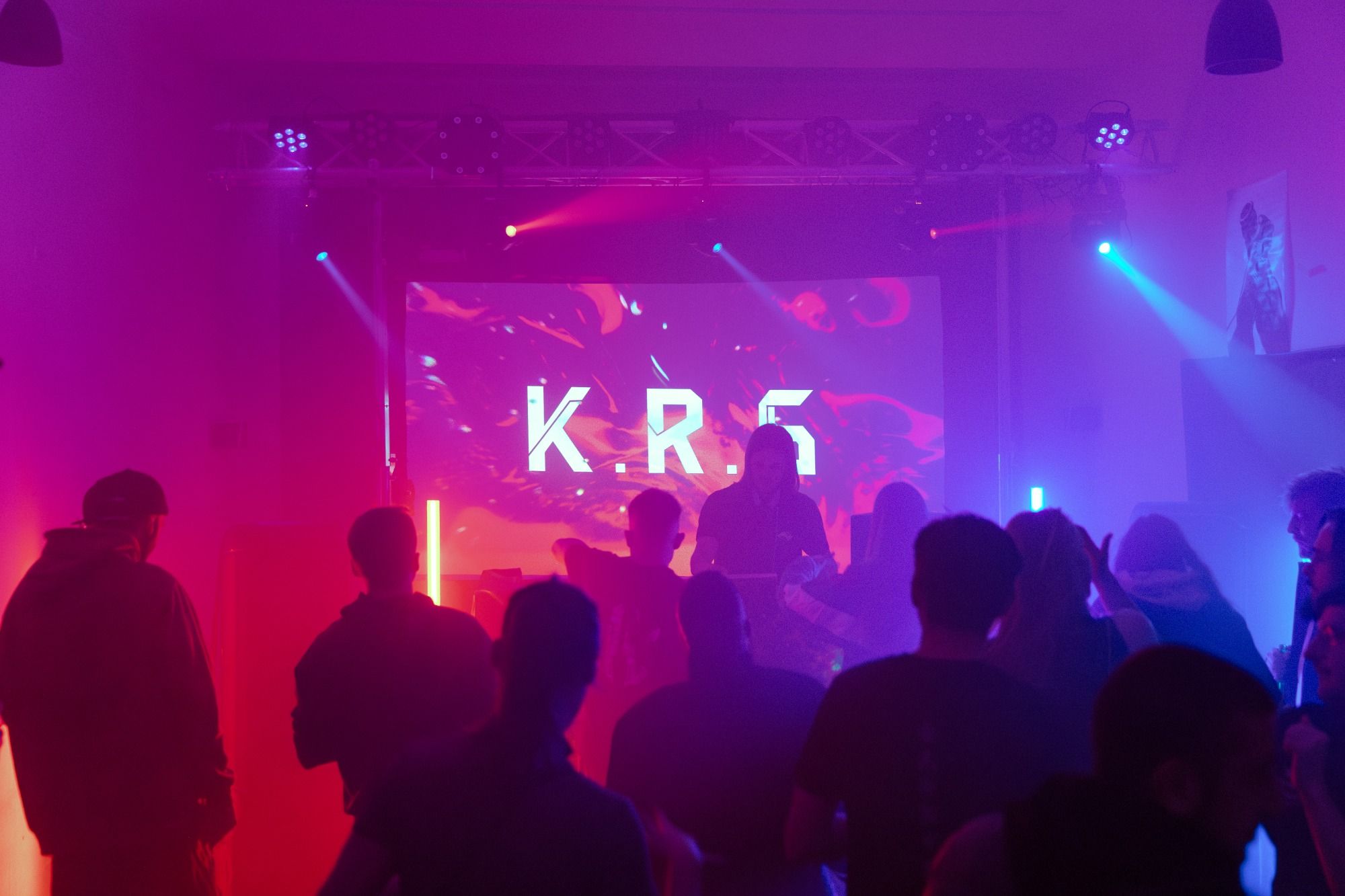 DJ za pultem před plátnem se jménem, dav ve fialovém a růžovém světle